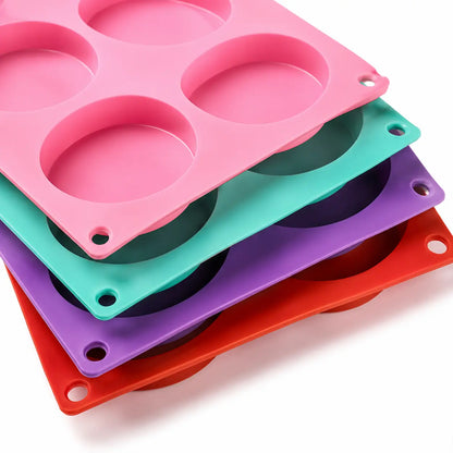 Four silicone mold trays with six circular cavities, stacked diagonally on a white background. The colors of the trays are pink, teal, purple, and red.